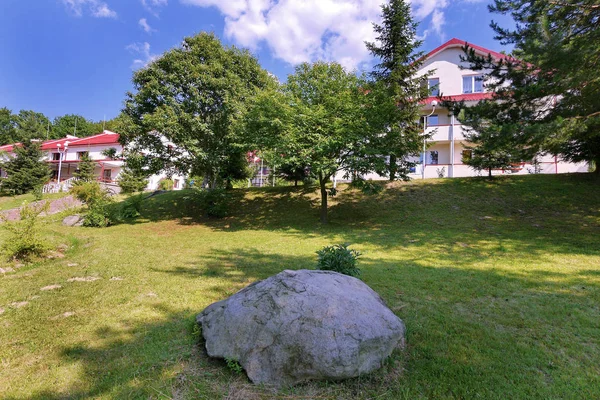Una gran piedra yaciendo en una pendiente verde que va detrás de las casas de pie sobre una colina contra un hermoso cielo azul . — Foto de Stock