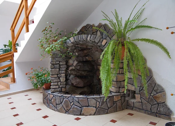 Decorative fireplace laid out of stones of different shapes in the room near the wall with pots standing on it with flowers.
