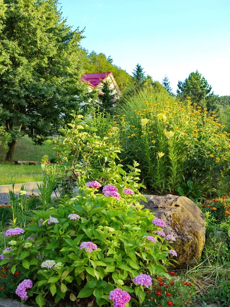 Большой камень, лежащий в клумбе с цветами, растущими на открытом воздухе рядом с зеленой лужайкой и деревьями . — стоковое фото