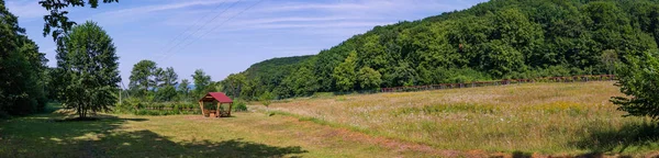 A panorama of meadow with a gazebo on the background of green wooded mountains on a clear day — Stock Photo, Image