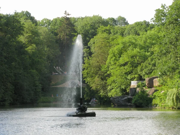 Ein hoher Springbrunnen, der in der Mitte der Wasseroberfläche des Teiches plätschert, mit Touristen am Ufer und einer Aussichtsplattform auf riesigen Felsbrocken. — Stockfoto