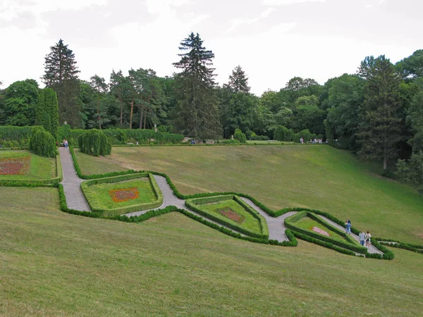Un hermoso paisaje de un césped verde en un parque en una pendiente con senderos que van entre macizos de flores figurados con personas que caminan por ellos . — Foto de Stock