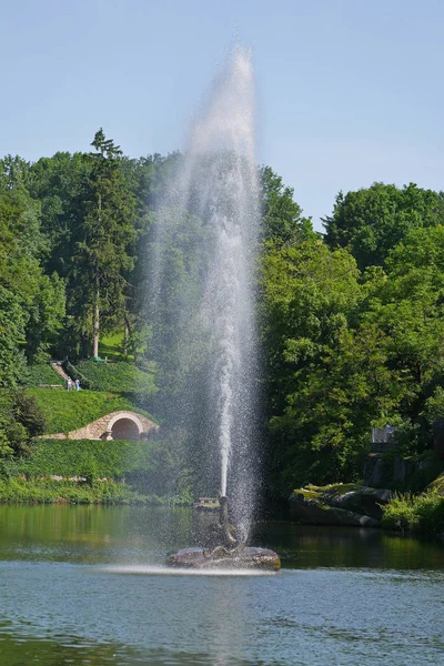 Öppet vattenfontän slående i mitten av sjön mot en bakgrund av grön natur och backar i parken med människor som vandrar längs stigar. — Stockfoto