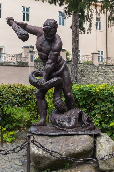 Escultura representando um homem com um baú de uma serpente conquistadora perto de seus pés. Em pé sobre as pedras atrás da cerca com uma corrente perto dos arbustos verdes na frente da casa . — Fotografia de Stock