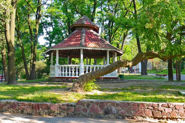 Laube in einem Park mit einem schrägen Dach steht im Schatten von Bäumen mit grünem Gras herum. ein guter Ort zum Entspannen im Freien. — Stockfoto