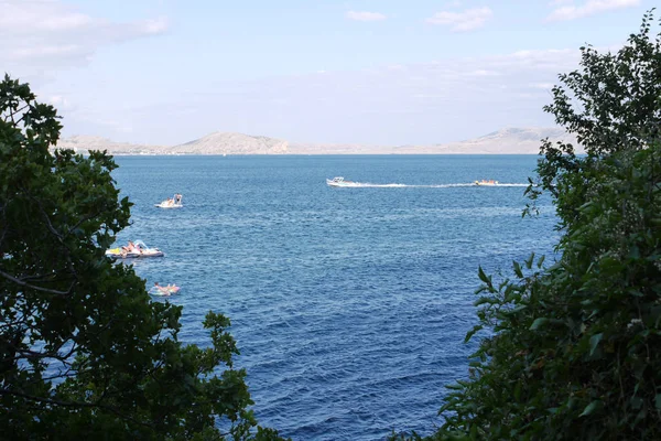 Blue view of the sea between green bushes. In the water people rest on catamarans, boats and boats