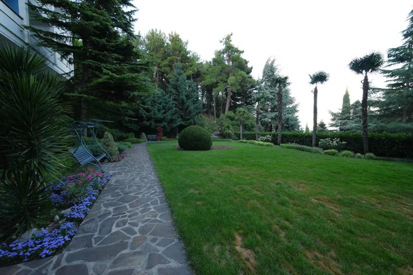 Un parque con un camino bordeado de piedra está plantado con varias plantas ornamentales bajo las ventanas de la casa . — Foto de Stock