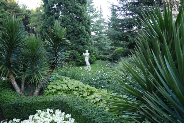 Gyönyörű szelíd virágok a parkban növekvő közepette buja növényzet és magas fák egy szobor gipszkarton mellett egy zöld gyep. — Stock Fotó