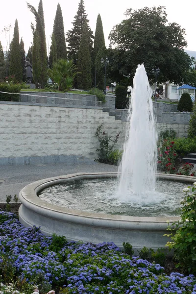 Een prachtige fontein met helder water met een prachtig bloembed met bloemen op de achtergrond van een monument dat in de verte tussen de hoge weelderige bomen staat. — Stockfoto