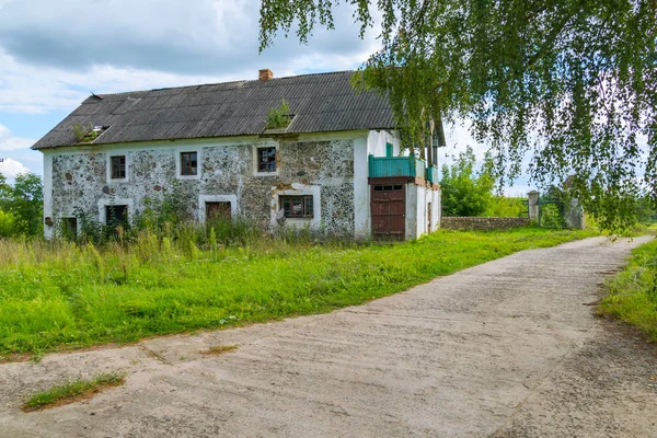 En lång bred väg är omgiven av gräs på bakgrunden av övergivna hus och höga, lummiga träd — Stockfoto