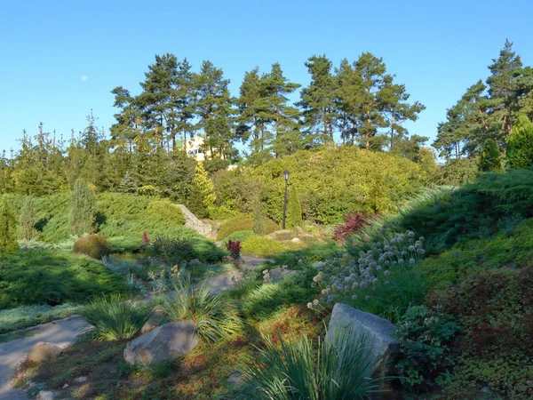 Parc vert avec un sentier allant entre les parterres de fleurs avec de magnifiques fleurs et de grandes pierres couchées en eux. Pelouses vertes et grands arbres . — Photo