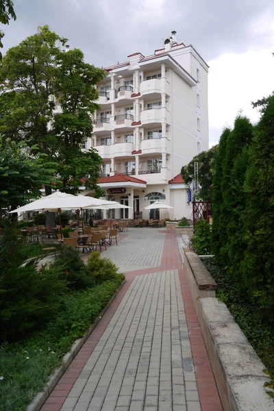 Der Weg zwischen grünen Büschen und Rasen führt zum Hotel mit Balkonen, daneben stehen Tische und Stühle für Touristen auf der Straße. — Stockfoto