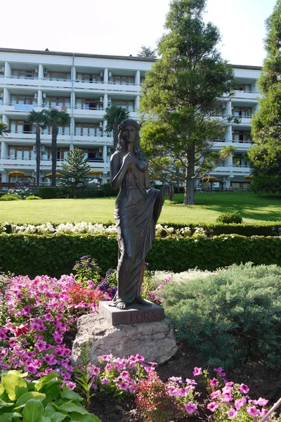 一位妇女在绿色的草坪上的雕塑, 站在一块石头上, 在弗洛拉的基座上刻有铭文, 酒店旁边有越来越多的鲜花, 有阳台. — 图库照片