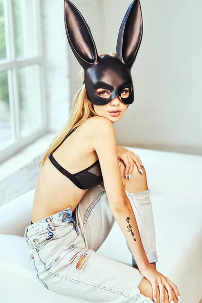 A model in a rabbit mask poses in a studio with a white background. Beautiful underwear in black, jeans and mask. Blonde with a beautiful figure