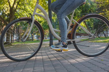 Kot pantolonlu ve spor ayakkabılı bir kız parkta yeşil ağaçların ve çimenlerin arasında bej bir bisiklete biniyor.
