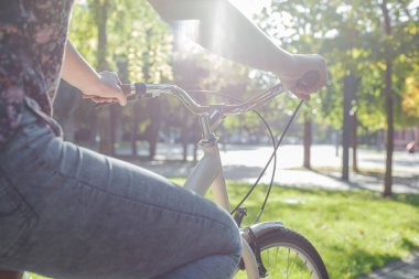 Kot pantolonlu ve spor ayakkabılı bir kız parkta yeşil ağaçların ve çimenlerin arasında bej bir bisiklete biniyor.