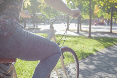 Kot pantolonlu ve spor ayakkabılı bir kız parkta yeşil ağaçların ve çimenlerin arasında bej bir bisiklete biniyor.