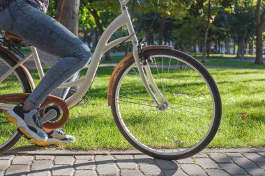 Kot pantolonlu ve spor ayakkabılı bir kız parkta yeşil ağaçların ve çimenlerin arasında bej bir bisiklete biniyor.