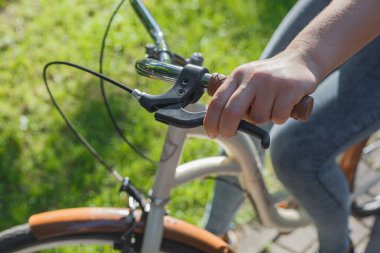 Bir kız bisikletin direksiyonunu tutuyor ve yeşil ağaçların ve çimlerin arasındaki bir parkta bej bir el freni tutuyor.