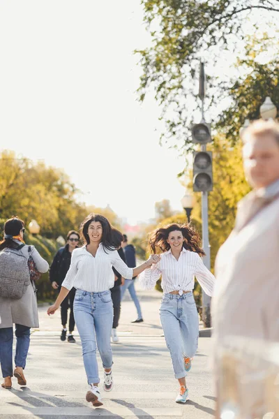 Neşeli iki kız arkadaş şehrin yaya geçidinde karşıdan karşıya geçerken kucaklaşırken eğleniyorlar.