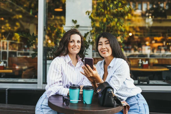 Gülümseyen neşeli kadın arkadaşlar kahve içiyorlar telefonla kameraya bakıyorlar caddedeki kafede oturuyorlar.