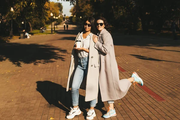 Ellerinde kahveyle sonbahar parkında yürüyen gözlüklü ve paltolu iki kadın.