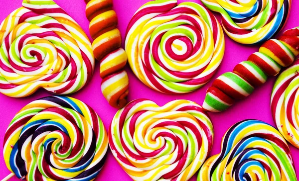 Rainbow lollipop swirl on wooden stick on pink paper background, flat lay