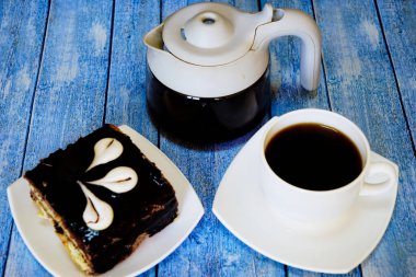 Beyaz fincanda sade kahve ve mavi ahşap bir masanın üstünde kremalı kek..