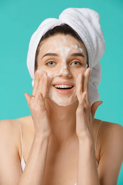Young woman with towel wrapped around her head applying facial cleaning foam and having fun. Daily routine - face cleaning, skin care, peeling, moisturising and beauty treatment concept