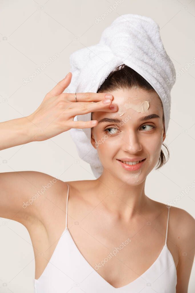 Young woman with towel wrapped around her body applying moisturing face cream and having fun. Daily morning routine - facial cleaning, skin care, peeling, moisturising and beauty treatment concept