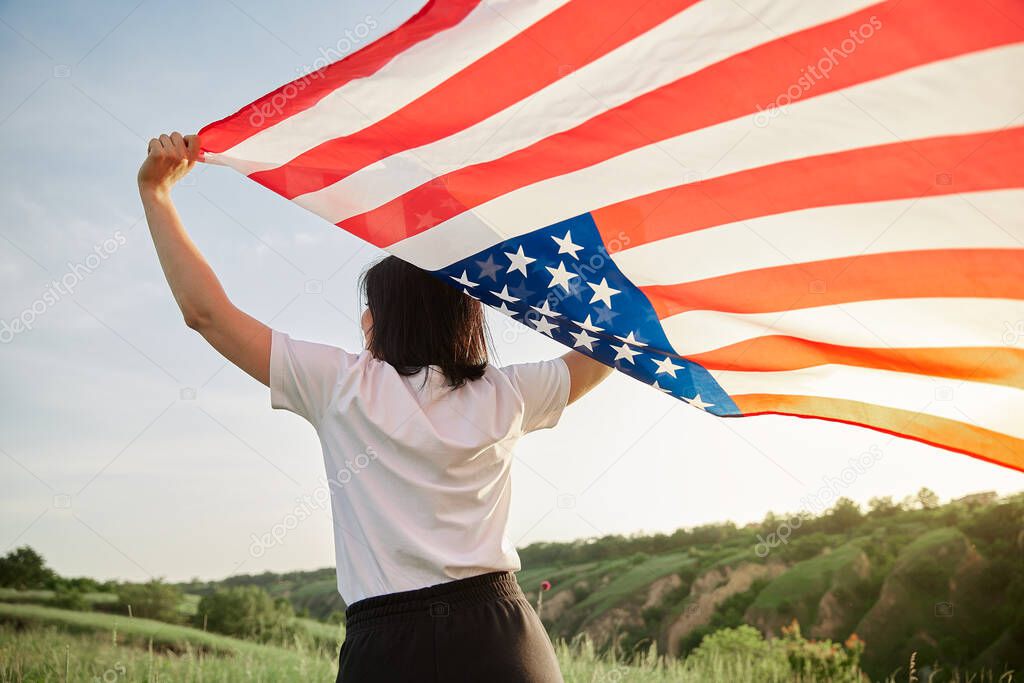 4th of July. Fourth of July. American woman with the national American flag against beautiful landmark. Independence Day. Beautiful sunset light. Patriotic holiday, democracy respect and veteran respect concept