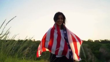 Rüzgarda dalgalanan Amerikan bayrağıyla sarmalayan kadın. 4 Temmuz, Bağımsızlık Günü kutlaması. Yavaş çekim geniş açı kamera takip ediyor. Ön manzara. Vatansever bayram, demokrasi ve gazi saygısı