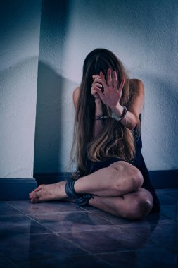 Genç bir kadın şiddete maruz kalıyor, yere oturuyor, ellerini kelepçeleyerek yüzünü kapatıyor. Elleri damarlar ve yaralar, bacakları bir iple bağlanmış. Travma sonrası stres bozukluğu.