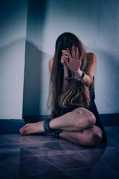 A young woman suffers from violence, sits on the floor, covers her face with hands in handcuffs. Her hands are veins and scars, her legs are tied with a rope. PTSD post-traumatic stress