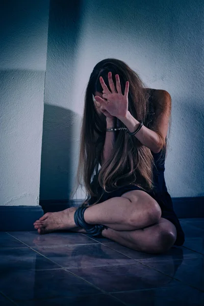 A young woman suffers from violence, sits on the floor, covers her face with hands in handcuffs. Her hands are veins and scars, her legs are tied with a rope. PTSD post-traumatic stress