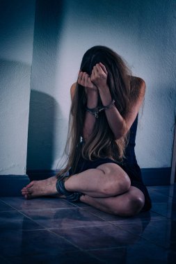 Genç bir kadın şiddete maruz kalıyor, yere oturuyor, ellerini kelepçeleyerek yüzünü kapatıyor. Elleri damarlar ve yaralar, bacakları bir iple bağlanmış. Travma sonrası stres bozukluğu.