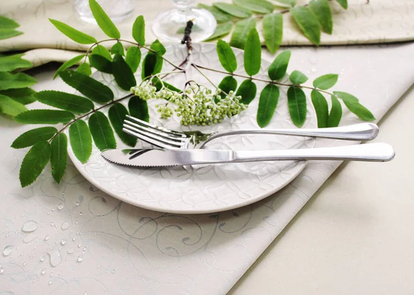 Masa düzenleme detayları. Masada bej tabaklar ve çatal bıçak seti. Dağ külünden yeşil bir dekoru var. Yukarıdan görünüm.