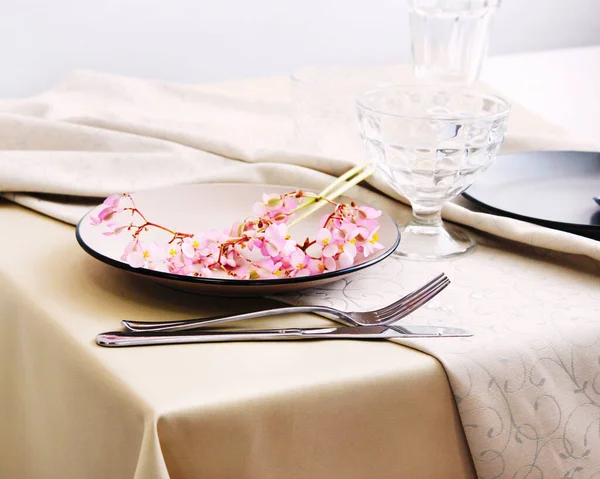 Masada pembe begonya çiçekleri dekoru olan bej tabaklar ve çatal bıçak takımı. İç kısımda servis masası var. tablo ayarı detayları.