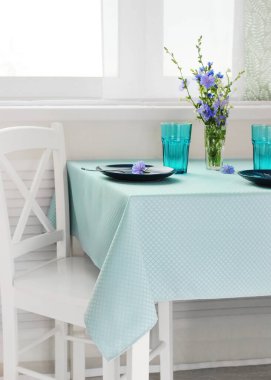 Masada mavi bir masa örtüsü ve parlak bir odada pencerenin yanında bulaşıklar. romantik yemek masası detayları ayarlıyor.