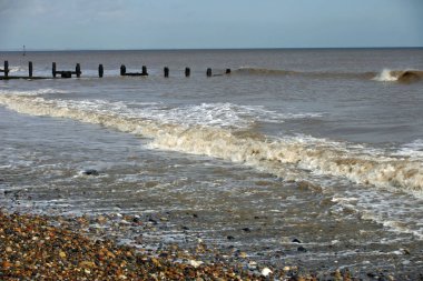 Ahşap dalgakıran (groyne) uzaktan çakıllı plaj sakin deniz kıyıya alıştırma ile ön planda. Arka plan olarak bulut beyaz ile mavi gökyüzü.