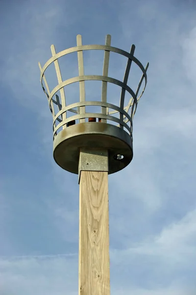 Beacon to be used with either a fire, gas or electric light. Metal basket construction. On a wooden post. Background of blue sky with white cloud.