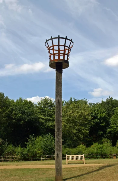 Fire beacon on a wooden pole set in lawns in a park with trees, bench and blue sky with white clouds behind.