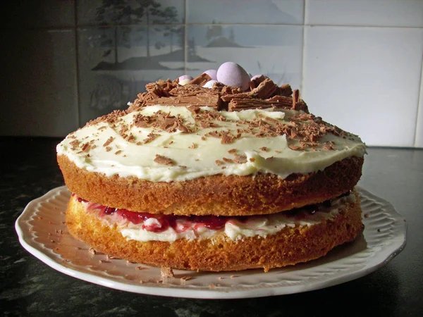 Home made Easter cake made of sponge with jam filling and chocolate flake and Easter egg topping. On a white plate. Lit from the side.  Background of dark granite coloured work surface and blue/grey tiles with pattern.