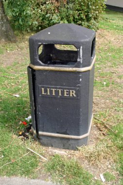 Siyah plastik çöp kutusu ile bazı çöp yanında yıpranmış. Biçilmiş ot ve çalılar arkasında çevrili.