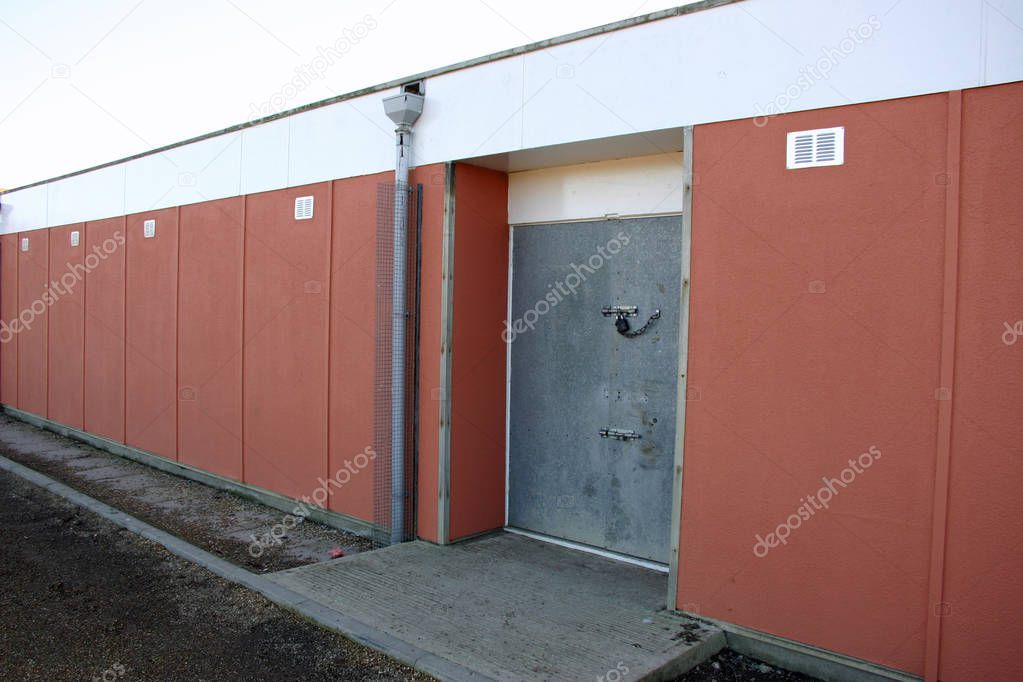 Secure double metal sports pavilion door with padlock and chain and concrete slope surrounded by a terracotta coloured wall with a pebbledash finish.