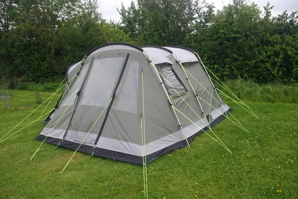 Grey and green four person tent with black sown-in groundsheet. Well guyed with bright yellow guy ropes. Erected on a grass area with trees and sky in the background.