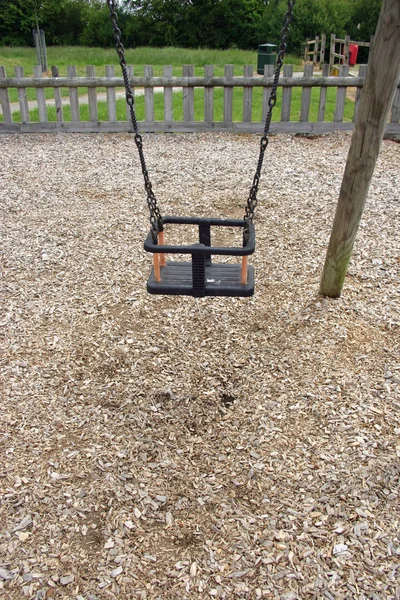 Park swing for a young child where the chipped bark is wearing away underneath. Fence, litter bin, grass and trees in the background.