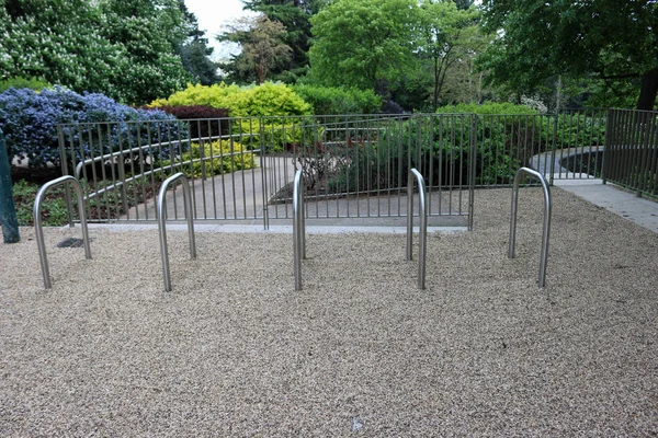 Five metal new bicycle racks on a gravel surfaced tarmac path in a park with a tree, shrub beds, fences and other park furniture behind.