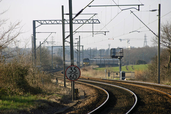 Curved local railway line with electric overhead cables and train in the distance. Background of trees, hedges and fields.