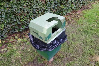 Zarar görmüş plastik kapak bir engel arkasında ve önünde bir çim çim ile tepesinde bir siyah plastik depo gözü liner ile eski ve yıpranmış yeşil boyalı metal çöp kutusu.